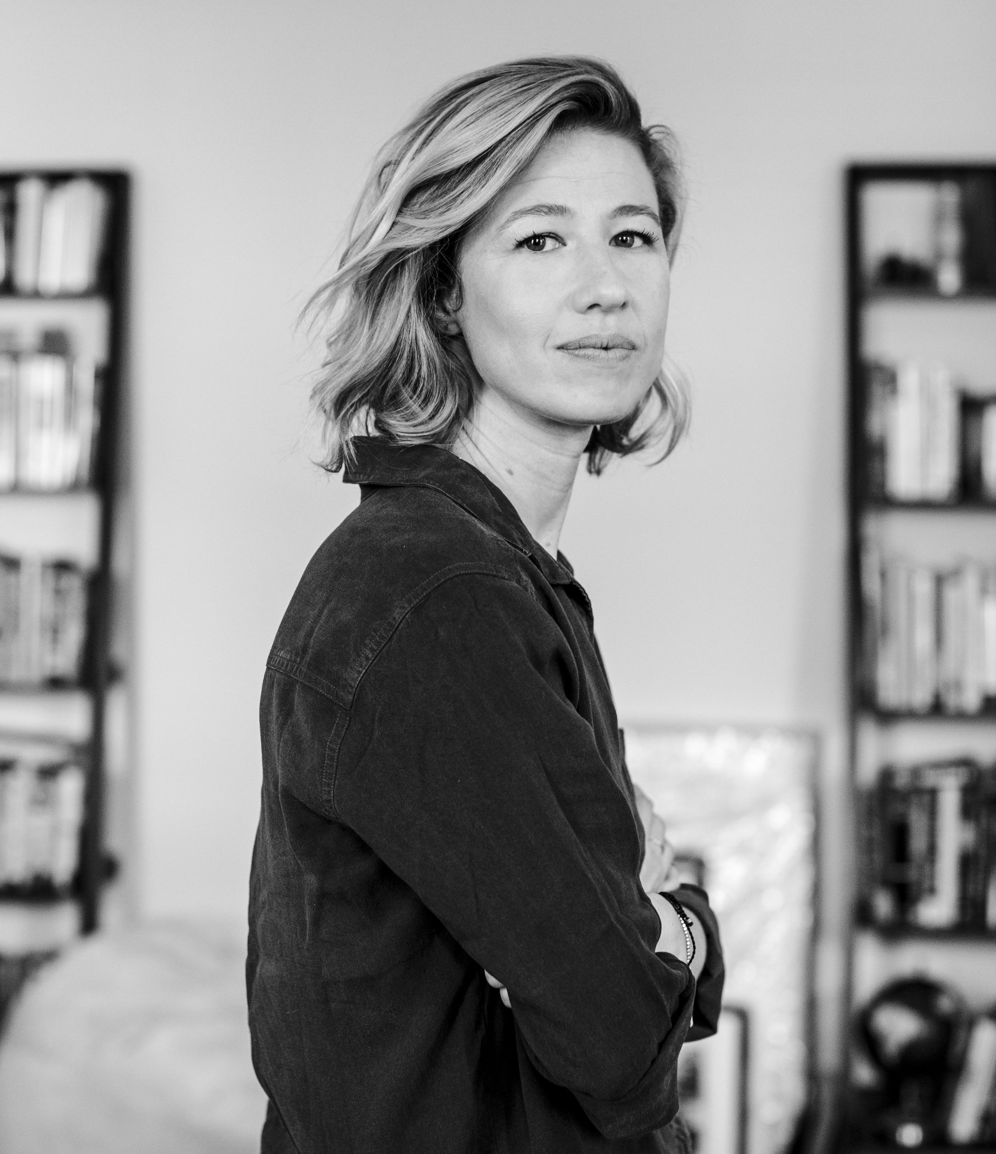 Photo of a white woman with shoulder-length hair, crossing her arms, looking at the camera with bookshelves in the background.