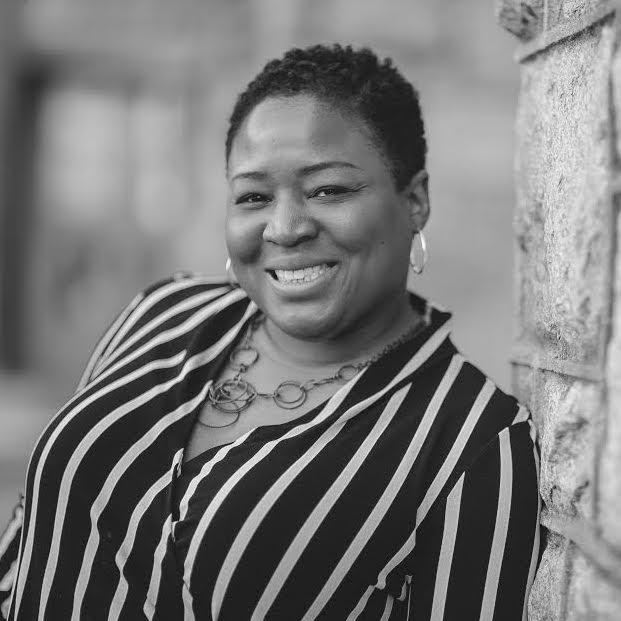 Jos Duncan-Asé looking at the camera, smiling. She is wearing a black and white vertical stripe blouse and hoop earrings, and is standing next to a stone wall. Black and white portrait, with the background out of focus.