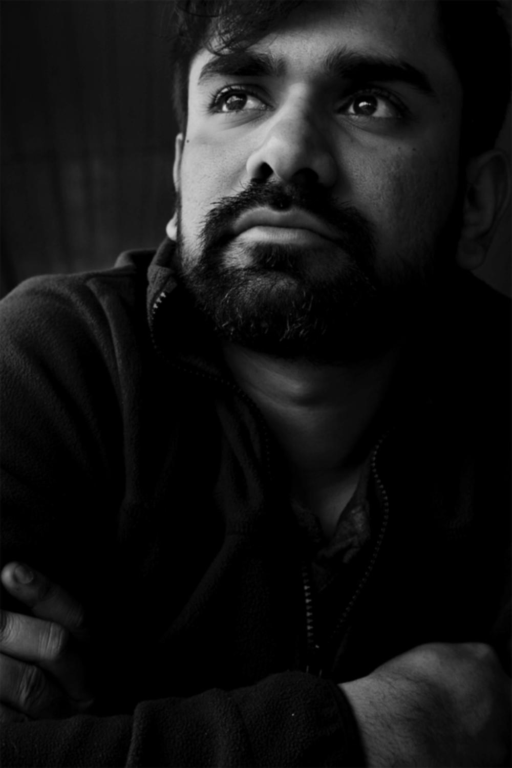 Headshot of an Indian man with black hair, wearing a black jacket and looking sideways against a dark gray background.